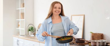 Mutfakta sebzeli kızartma tavası tutan genç bir kadın.