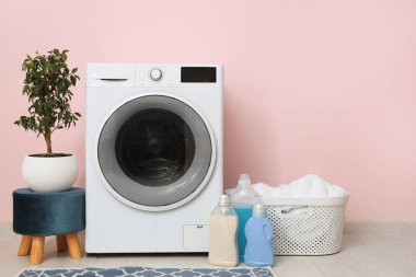 Pembe duvarın yanında sepetli, çiçekli ve deterjanlı modern çamaşır makinesi.