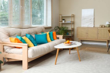 Şık oturma odasının içinde rahat bir kanepe, sepetin içinde kahve masası ve pencere var.