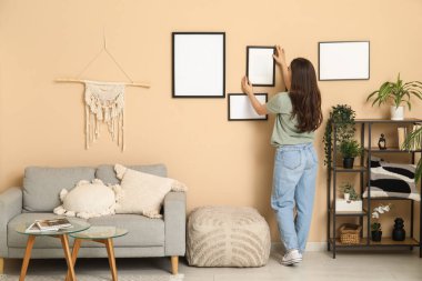 Oturma odasındaki bej duvarda boş bir çerçeve asılı genç bir kadın. Arka plan