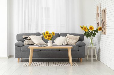 Rahat bir kanepe, tabure ve vazolar. Oturma odasının içinde güzel ayçiçekleri var.