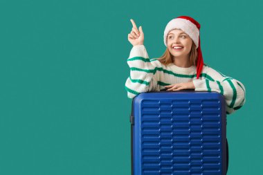 Yeşil arka planda işaret parmağı ve bavulu olan Noel Baba şapkalı bir kadın yolcu.