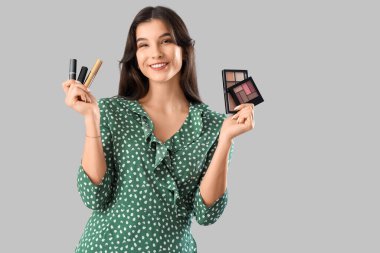 Gri arka planda farklı kozmetik ürünleri tutan güzel genç bir kadın.