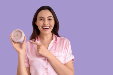 Pembe pijamalı genç bir kadın leylak arka planındaki çalar saati işaret ediyor.