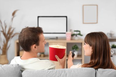 Patlamış mısırlı genç çift evdeki kanepede televizyon izliyor.