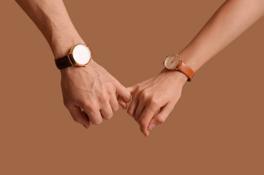 Kol saatleri olan çift kahverengi arka planda serçe parmak tutuyor.