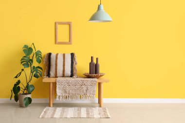 Sarı duvarın yanında bank, ev bitkisi ve yastıklı bir oda.