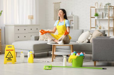 Lastik eldivenli genç kadın oturma odasındaki temizlik malzemelerinin arasında meditasyon yapıyor.