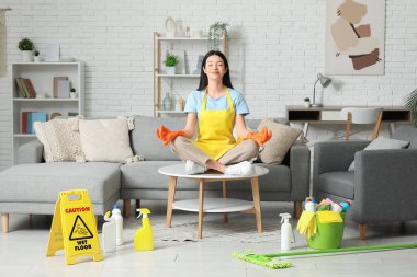 Lastik eldivenli genç kadın oturma odasındaki temizlik malzemelerinin arasında meditasyon yapıyor.
