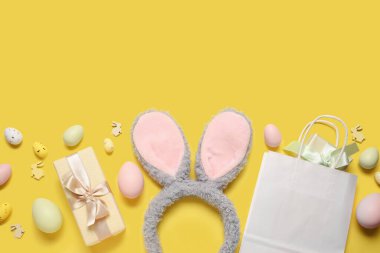 Tavşan kulaklı alışveriş çantası, hediye kutuları ve sarı arka planda Paskalya yumurtaları.