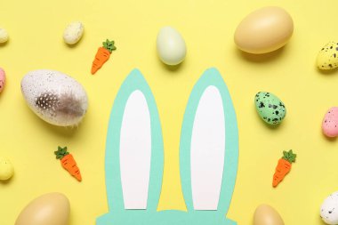 Paskalya yumurtaları ve sarı arka planda dekoratif havuçlarla kağıttan tavşan kulakları.