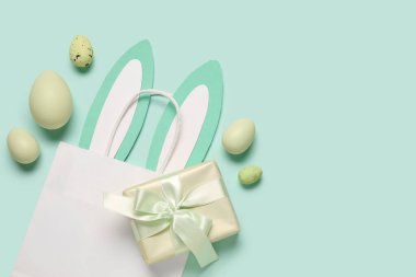 Kağıt tavşan kulaklı alışveriş torbası, Paskalya yumurtası ve turkuaz arka planda hediye kutusu.