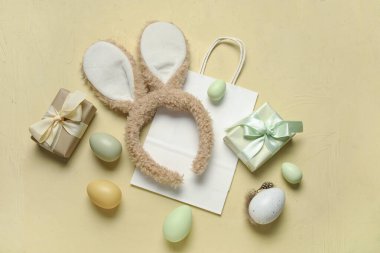Tavşan kulaklı alışveriş çantası, hediye kutuları ve sarı arka planda Paskalya yumurtaları.
