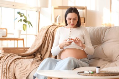 Genç hamile kadın evdeki kanepede glukometre kullanıyor. Gebelik Diyabet Konsepti