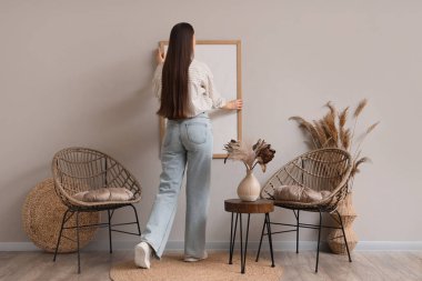 Odadaki gri duvara çerçevesini asan genç bir kadın. Arka plan