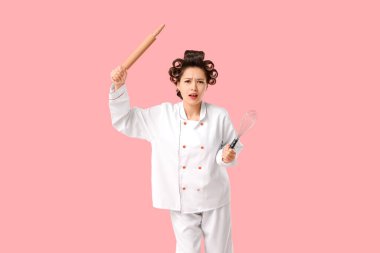 Pembe arka planda saç bigudileri ve mutfak gereçleri olan hoşnutsuz Asyalı kadın şef.