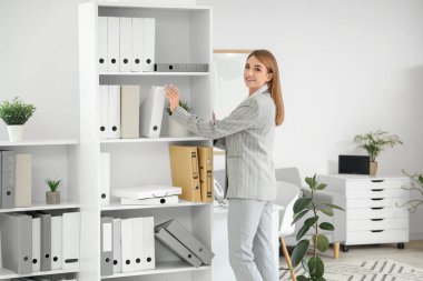 Genç iş kadını, ofisteki raftan evrak klasörünü alıyor.