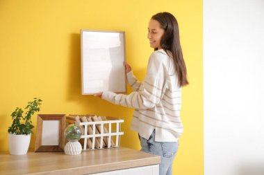 Genç bir kadın mutfaktaki sarı duvara boş bir çerçeve asıyor.