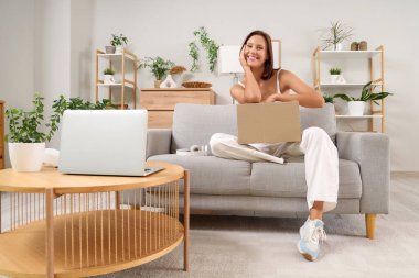 Evdeki kanepede oturan koli ve dizüstü bilgisayarı olan güzel, genç ve mutlu bir kadın.