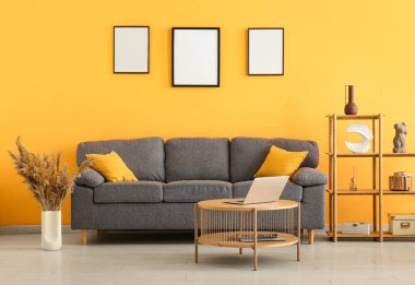 Koltuk, dizüstü bilgisayarlı masa ve sarı duvarda boş posterler.