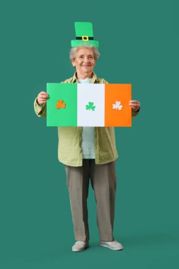 Yeşil arka planda Aziz Patrick Günü kutlamaları için İrlanda bayrağı taşıyan cüce şapkalı mutlu yaşlı kadın.