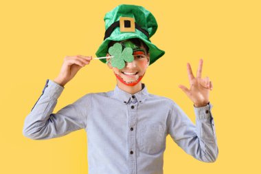 Yüz boyama, yonca ve cüce şapkalı komik çocuk sarı arka planda zafer jesti gösteriyor. Aziz Patrick Günü kutlaması