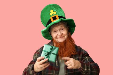 Kırmızı sakallı cüce şapkalı yaşlı kadın pembe arka plandaki hediye kutusunu işaret ediyor. Aziz Patrick Günü kutlaması