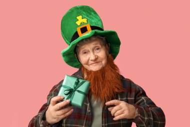Kırmızı sakallı cüce şapkalı yaşlı kadın pembe arka planda hediye kutusu tutuyor. Aziz Patrick Günü kutlaması