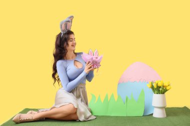 Tavşan kulaklı genç kadın. Paskalya pastası ve çimlerin üzerinde sarı arka planda dekor var.