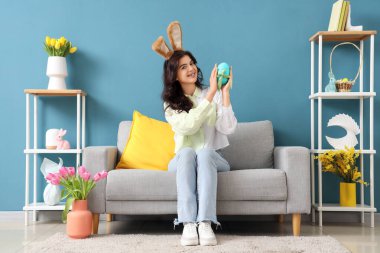 Tavşan kulaklı genç bir kadın. Paskalya yumurtası evde, kanepede oturuyor.