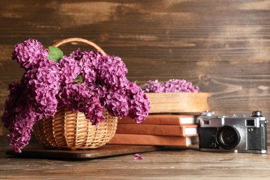 Tahta arka planda çiçek açan leylak çiçekleri, kitaplar ve fotoğraf makinesi olan sepet.