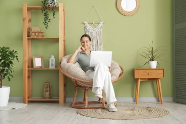 Genç bir kadın oturma odasındaki koltukta otururken dizüstü bilgisayar kullanıyor.