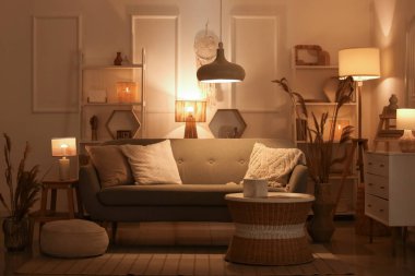 Rahat bir kanepe, çekmeceler, kahve masası, otlar ve oturma odasının içinde parlayan lambalar.