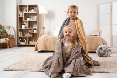 Küçük kız ve erkek kardeş evde kareli kumaşların altında yerde oturuyor.