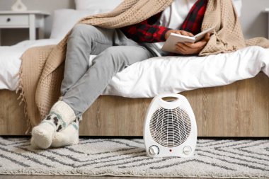 Yatak odasında elektrikli yelpaze ısıtıcısının yanında ekose kitap okuyan genç bir adam.