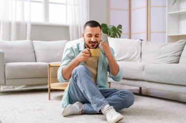 Seramik bardaklı genç adam evde yerde oturuyor.