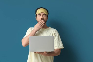 Uyku maskeli ve dizüstü bilgisayarı mavi arka planda esneyen uykulu genç adam.