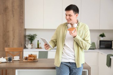 Genç adam elinde tablet bilgisayar ve tostla mutfakta fıstık ezmesi tutuyor.