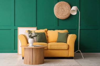 Konforlu sarı kanepe ve okaliptüs dalları olan kahve masası yeşil duvarın yanındaki vazoda.