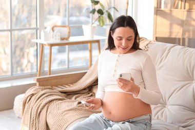 Genç hamile bir kadın evdeki koltukta glukometreyle cep telefonu kullanıyor. Gebelik Diyabet Konsepti