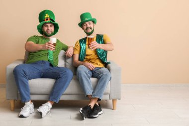 Yeşil sakallı, leprikon şapkalı, elinde bira bardakları olan ve bej duvarın yanındaki kanepede oturan mutlu genç adamlar. Aziz Patrick Günü kutlaması