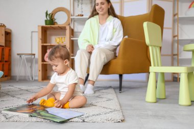 Kitabı halıya oturmuş, annesi evde olan tatlı bir kız.