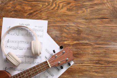 Ukulele, müzik kağıtları ve ahşap arka planda modern kulaklıklar.