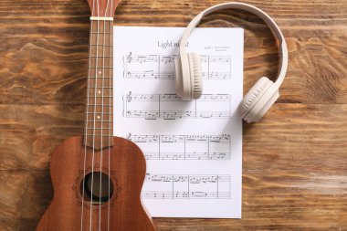 Ukulele, müzik sayfası ve ahşap arka planda modern kulaklıklar.