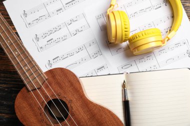 Ukulele, modern kulaklıklar, defter ve nota kağıtları ahşap masada, yakın plan.
