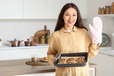 Genç Asyalı kadın mutfakta kurabiye tepsisi ve fırın eldiveni tutuyor.