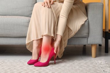 Young woman feeling pain in ankle because of uncomfortable high heeled shoes at home clipart