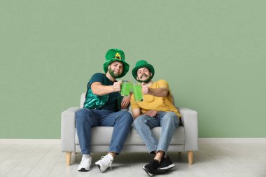 Yeşil sakallı ve yeşil sakallı mutlu genç adamlar ellerinde bira bardakları ve yeşil duvarın yanındaki kanepede oturuyorlar. Aziz Patrick Günü kutlaması