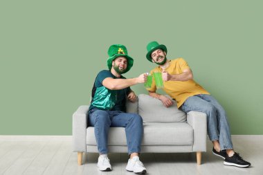 Yeşil sakallı ve yeşil sakallı mutlu genç adamlar ellerinde bira bardakları ve yeşil duvarın yanındaki kanepede oturuyorlar. Aziz Patrick Günü kutlaması