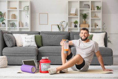 Evde yoga minderinde oturmuş protein içeceği içen sportif genç bir adam.
